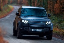 Land Rover Defender 130 V8 review: front three quarter driving, British B-road, tree-lined, low angle, black paint