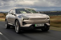Image of a beige Polestar 3, driving along a straight country road, viewed from the front right three-quarters