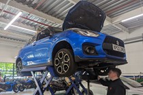 Suzuki Swift Sport (2023) long term test: front three quarter static, on the hoist in Suzuki's workshop for its first service, bonnet up, blue car