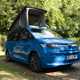Image of a blue Volkswagen California pitch at a wooded campsite with roof raised, viewed from the front right corner