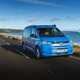 Image of a blue Volkswagen California driving along a country road, viewed from the front right corner