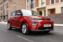 Kia Soul EV (2023): front three quarter tracking driving, orange paint, buildings in background