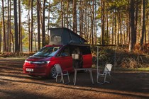 Volkswagen of a Volkswagen California parked in the woods, with pop top raised, awning and furniture out