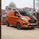 Image of an orange Ford Transit Custom, static in a builders merchant yard, viewed from the front right corner