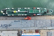 Image of a car transport ship moored at a dock, with ranks of cars waiting to be loaded