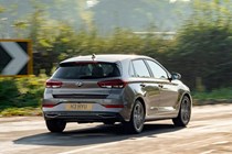 Hyundai i30 (2022) review - rear cornering shot, corner exit, brown car, leafy road, sign in background