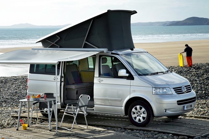 Image of Volkswagen California T5, in silver, with roof raised, boot open, and awning out with table and chairs set upImage of Volkswagen California T5, in silver, with roof raised, boot open, and awning out with table and chairs set up, from front