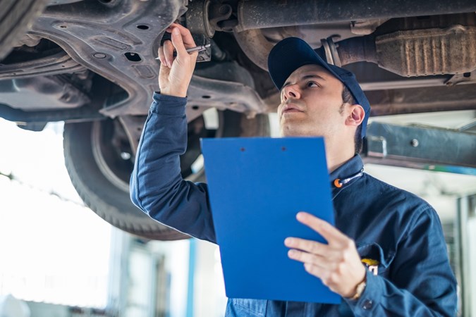 A mechanic is checking the underbody of a vehicle, holding a clipboard with an inspection sheet.