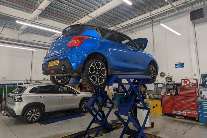 Suzuki Swift Sport (2023) long term test: rear three quarter static, on the hoist in Suzuki's workshop for its first service, bonnet up, blue car