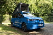 Image of a blue Volkswagen California pitch at a wooded campsite with roof raised, viewed from the front right corner