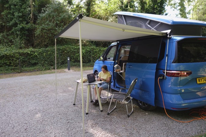 Image of woman relaxing under awning of Volkswagen California pitched at wooded campsite