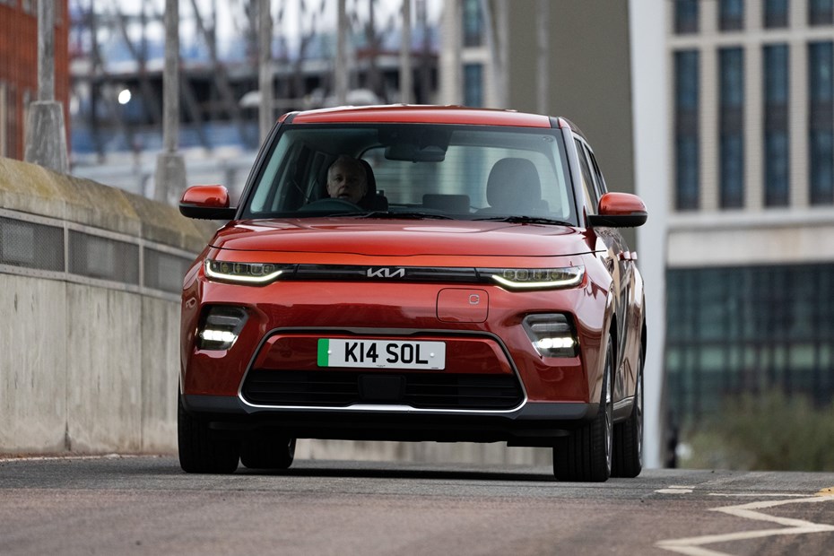 Kia Soul EV (2023): front three quarter driving, orange paint, buildings in background