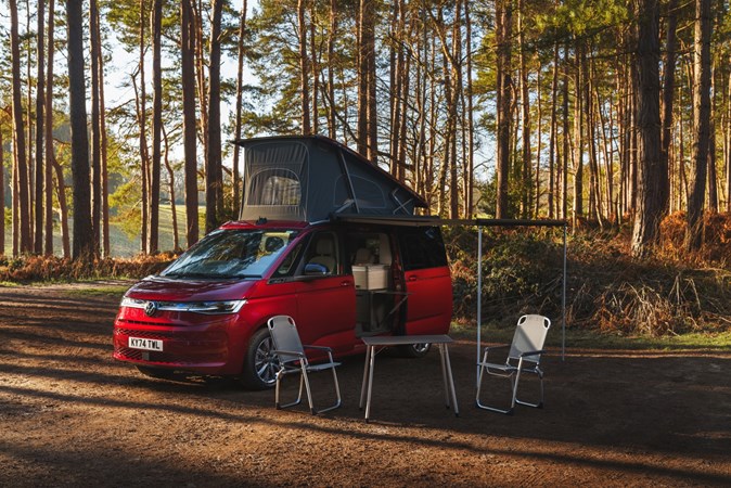 Volkswagen of a Volkswagen California parked in the woods, with pop top raised, awning and furniture out
