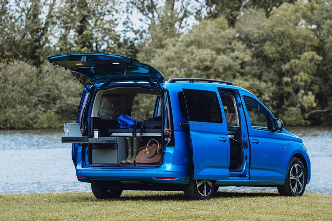 Image of a Volkswagen Caddy California parked on grass, seen from the rear right three-quarters, with tailgate open and kitchenette pulled out