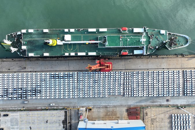Image of a car transport ship moored at a dock, with ranks of cars waiting to be loaded